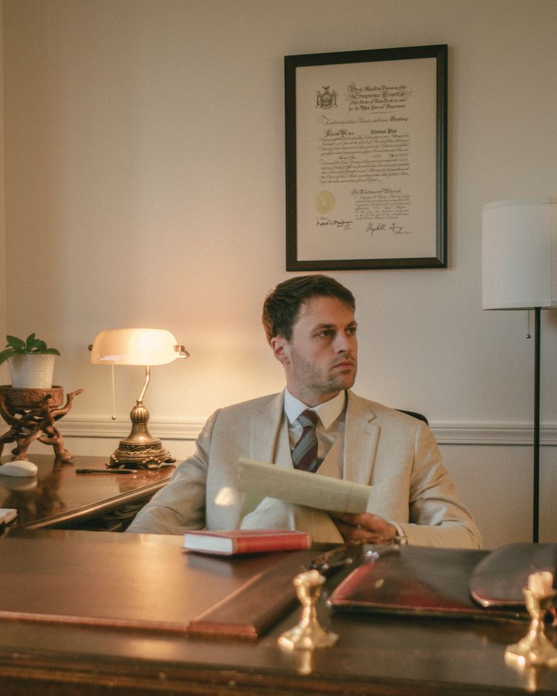 Thomas Plan am Schreibtisch, ein Schriftstück in der Hand, den Blick zur Seite; an der Wand hinter ihm hängt gerahmt die Urkunde seiner Zulassung in New York.